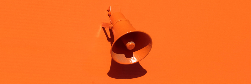 Slika prikazuje megafon, ki je pritrjen na oranžni steni